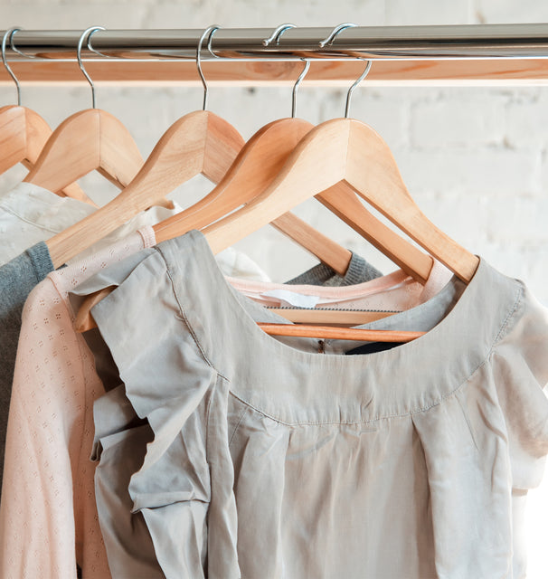Photo. Women's shirts on wooden hangers, hanging on clothing rack. Light pastel colored clothing against a white painted brick wall.